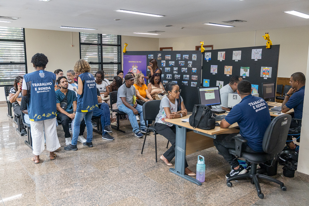 Imagem de grupo de pessoas sentadas em cadeiras sendo atendidas por funcionários da Secretaria Municipal de Trabalho e Renda uniformizados com coletes azuis com frase "Trabalho e Renda"