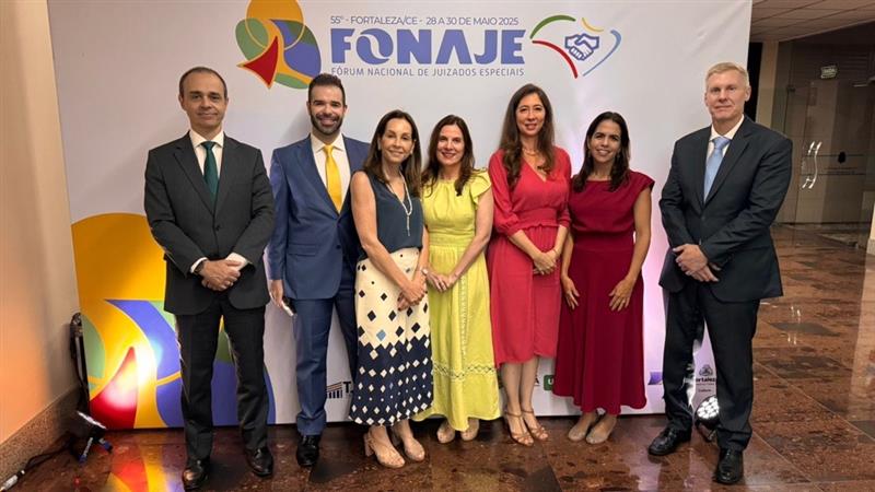 Foto mostra sete pessoas de pé, sendo dois homens de terno à esquerda, quatro mulheres de vestidos de cores diferenciadas no centro, e um homem de terno à direita. Todos são magistrados do TJRJ e posam à frente de painel de fundo branco com o símbolo colorido e a palavra Fonaje escrita com  letras grandes 