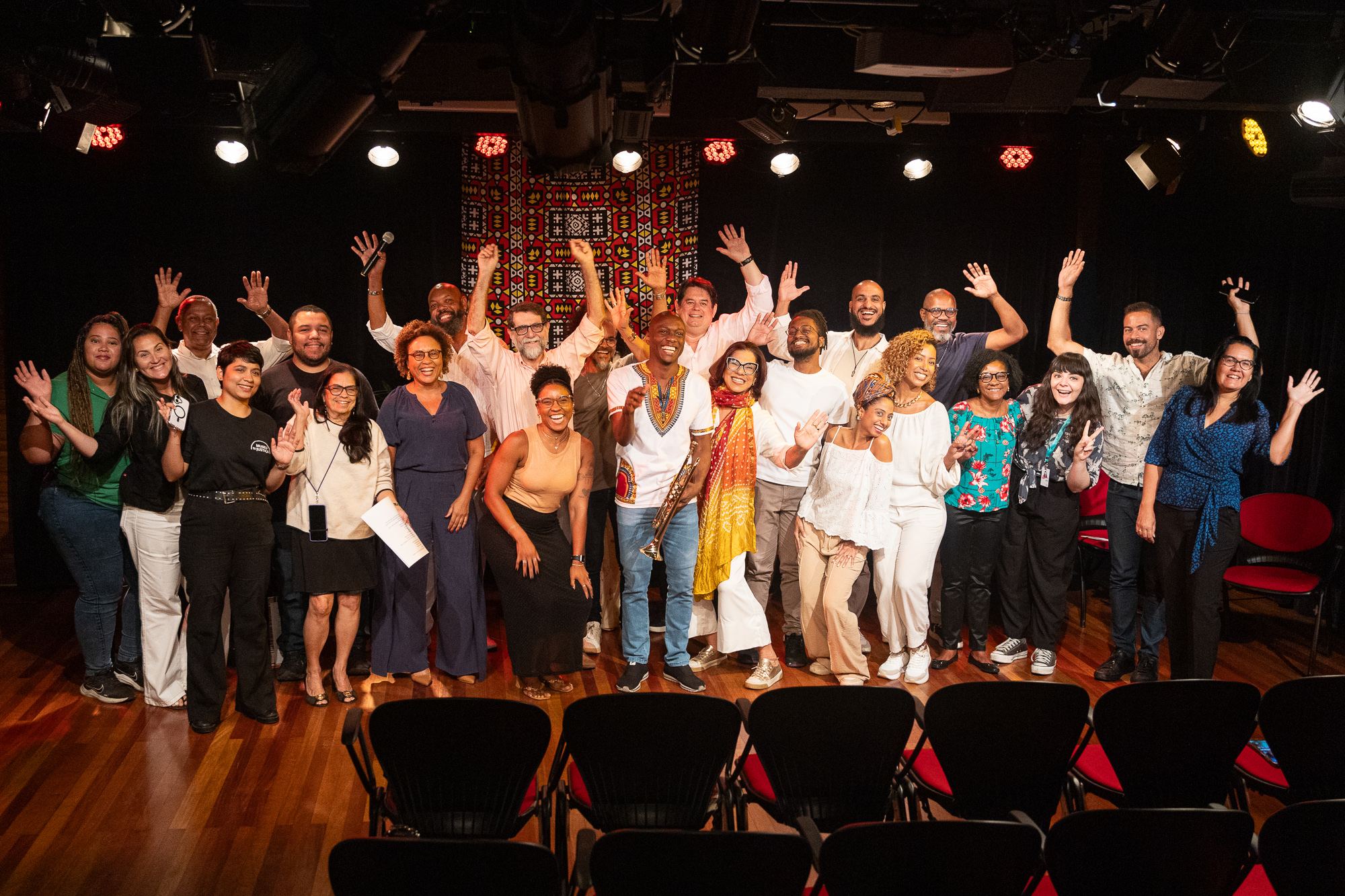 Imagem de grupos de pessoas no palco da Sala Multiuso sorrindo e com os braços para o alto