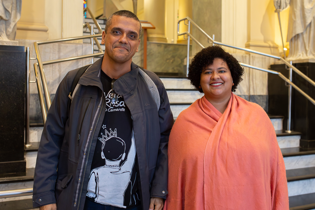 A imagem mostra duas pessoas posando lado a lado. Elas estão em frente a uma escadaria de mármore, com corrimãos metálicos e elementos arquitetônicos clássicos ao fundo, como colunas e iluminação suave, que valorizam o espaço. À esquerda, um homem veste jaqueta escura sobre uma camiseta com estampa gráfica e carrega uma mochila, mantendo uma expressão serena. À direita, uma mulher sorri para a câmera, usando um xale ou echarpe em tom coral, que se destaca no conjunto da imagem. 