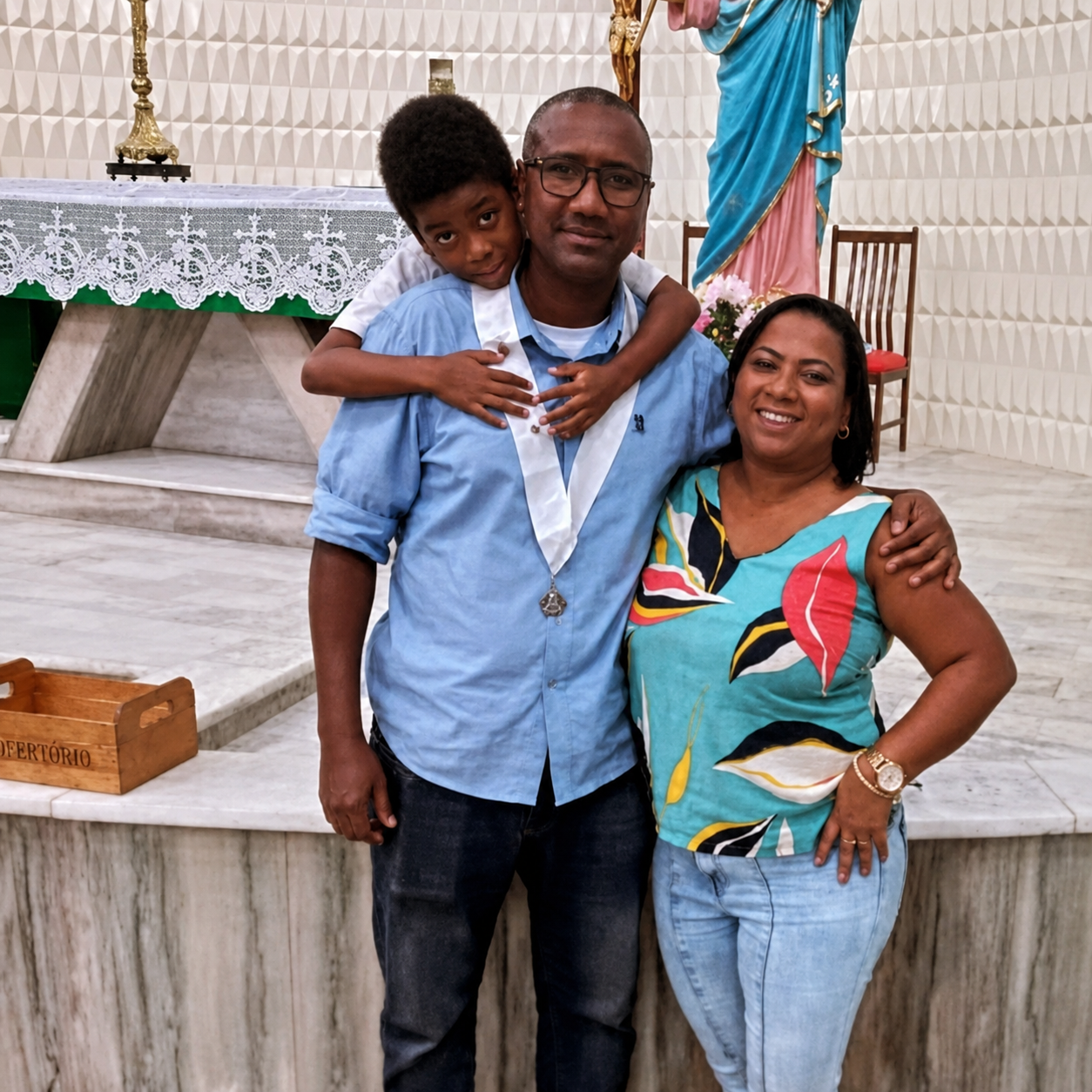 Imagem mostra uma família - um homem, uma mulher e um menino abraçando o homem pelas costas - em cenário de uma sacristia em tons claros