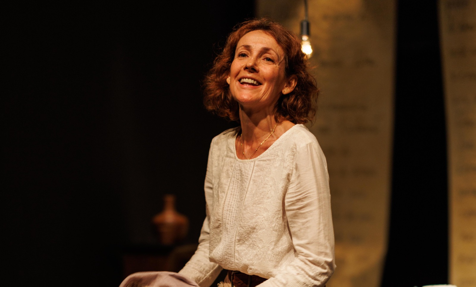 Atriz Ana Barroso em cena de palco, iluminado com luz amarelada e fundo escuro. Ela veste roupas que remetem ao século XVIII, saia comprida e blusa de mangas longas