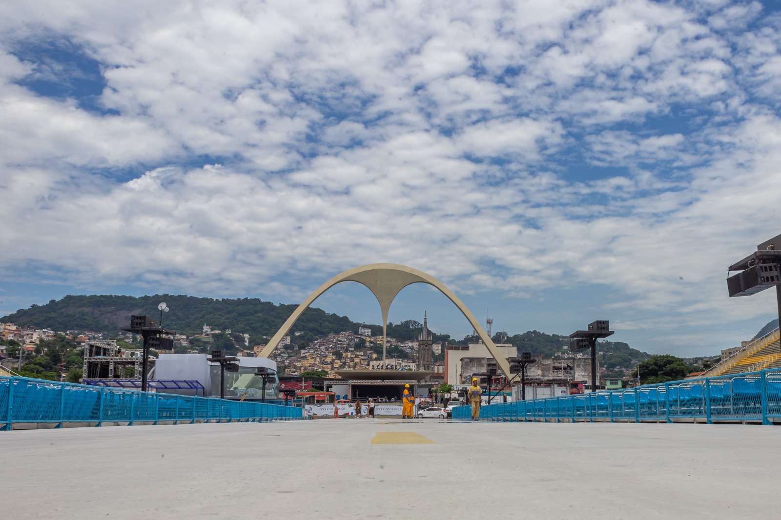 Foto do sambódromo do Rio, tirada do meio da pista, com cercas laterais azuis e com o símbolo da apoteose no fundo 