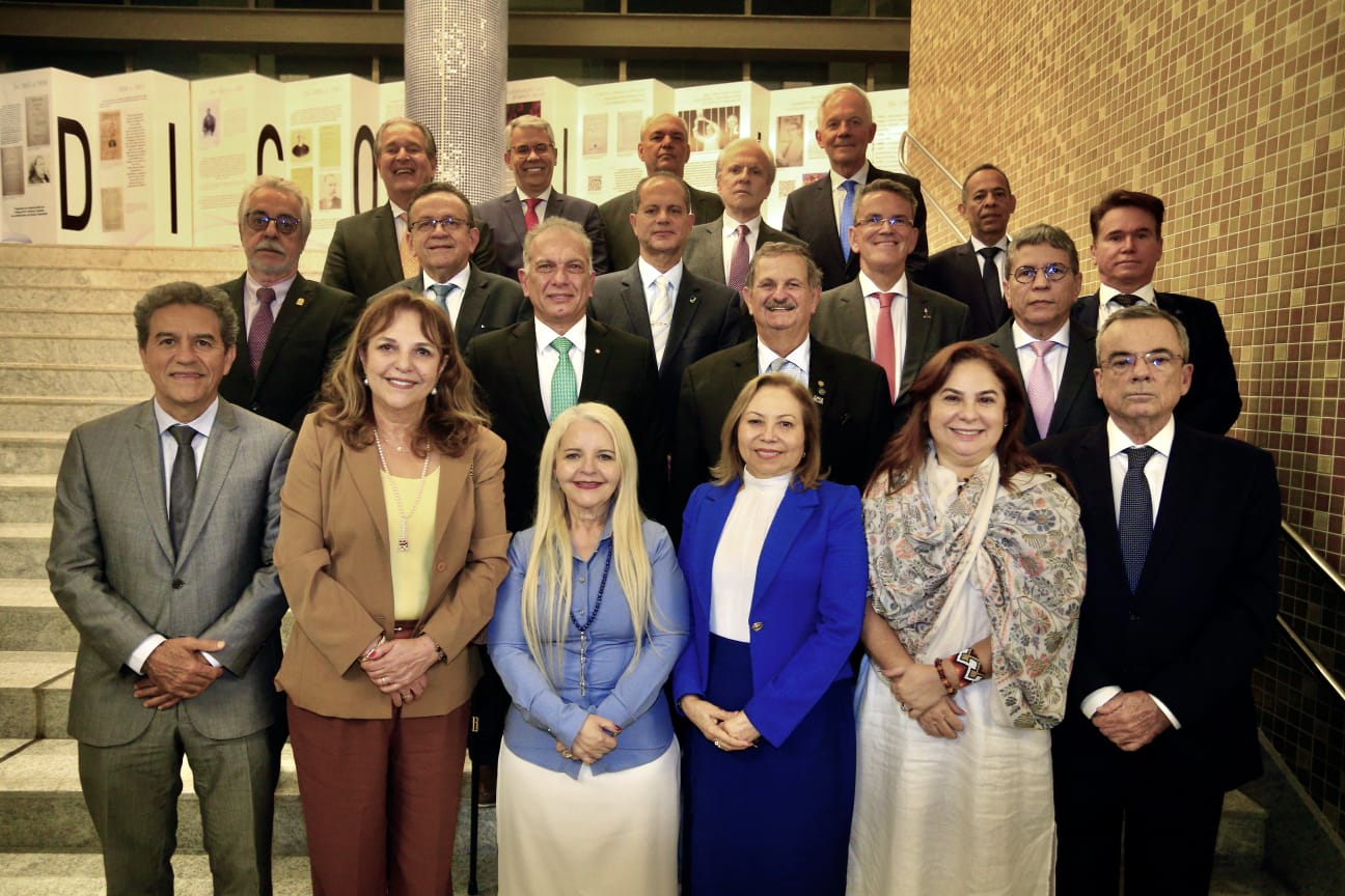 A imagem mostra um grupo de autoridades posando para uma foto institucional em um ambiente interno. As pessoas estão organizadas em quatro fileiras sobre uma escadaria, formando uma composição típica de retrato oficial. A maioria veste trajes formais, como ternos escuros, gravatas e blazers, enquanto algumas mulheres usam blazers, vestidos e lenços, também em estilo formal. Muitas das pessoas apresentam expressões sorridentes ou neutras, olhando diretamente para a câmera.