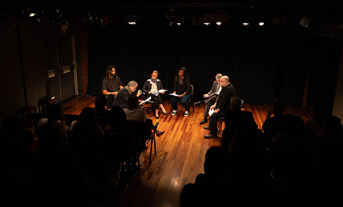 A imagem mostra seis pessoas (duas mulheres e quatro homens) sentados em cadeiras no centro do palco da Sala Multiuso e lendo textos.