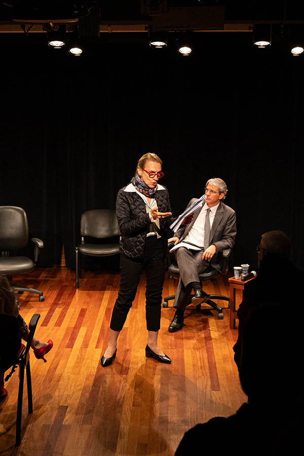 A imagem mostra a desembargadora Cristina Gaulia em pé lendo um texto no centro do palco da Sala Multiuso. Ao fundo, sentado em uma cadeira no palco, está o advogado José Roberto de Castro Neves. 