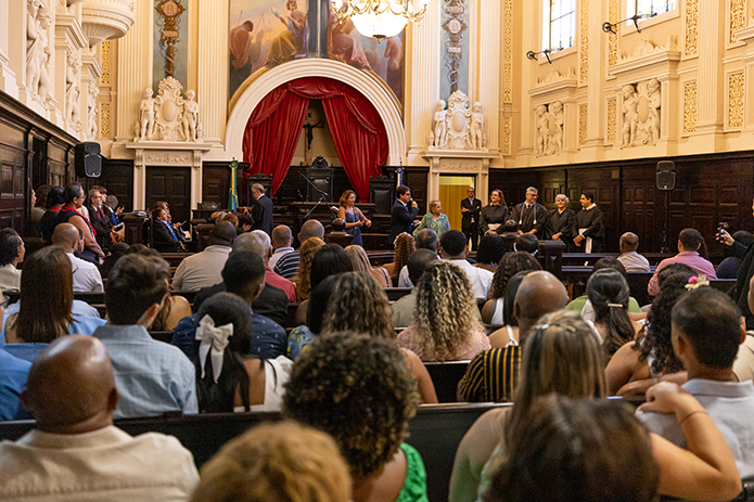 A imagem mostra o plenário do antigo Tribunal do Júri com juízes e servidores ao fundo e a plateia lotada de casais.