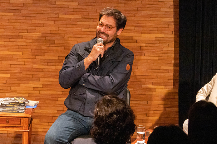 O ator Thiago Lacerda declama poesias sentado em uma cadeira no palco da Sala Multiuso