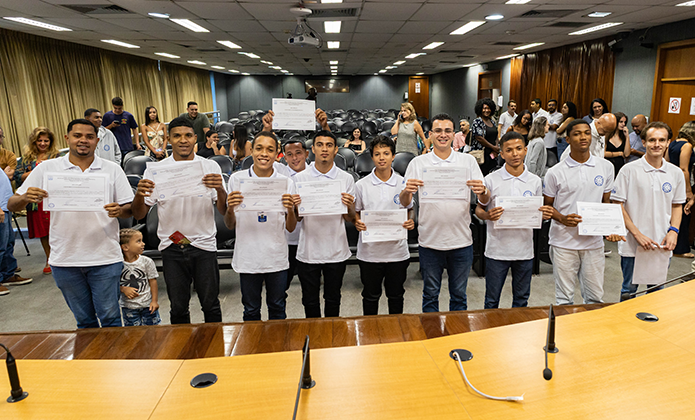Jovens, de camisas brancas e calcas jeans posam para foto junto ao palco de um auditório segurando seus diplomas