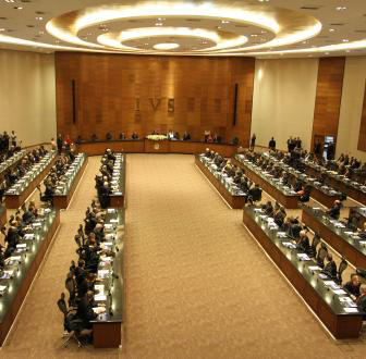 Sala de reuniões com várias mesas dispostas em fileiras, onde diversas pessoas estão sentadas, aparentemente em uma sessão deliberativa.