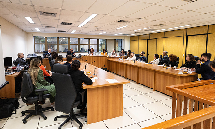 Foto de reunião, em sala de piso claro, paredes claras e janela ampla, com a presença de pelo menos 23 pessoas, todas sentadas em mesas de madeira posicionadas em formato de U e tendo à cabeceira uma mulher na cadeira mais alta , e uma mulher a sua direita e um homem a sua esquerda. Sobre as mesas copos de água e xícaras de cafés, além de microfones