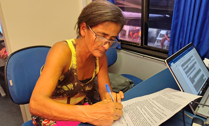 Foto mostra mulher de cabelos grisalhos, de óculos, sentada dentro do ônibus da Justiça Itinerante, assinando um documento sobre mesa diante de um computador. portátil