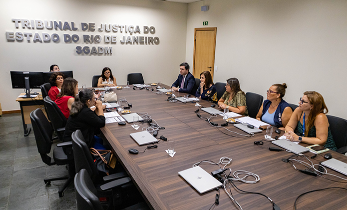 Reunião com dez pessoas em sala da Secretaria Geral de Administração, cujo letreiro aparece em média dimensão em uma das paredes brancas da sala. Ao redor de uma grande mesa de tampo na cor marrom e com notebooks sobre ela estão os participantes da reunião, sendo nove mulheres e um homem