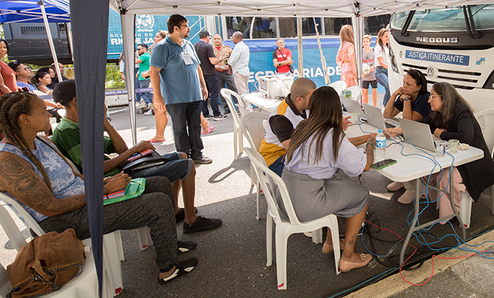 Foto mostra pessoas sendo atendidas por integrantes do Programa Justiça Itinerante em área externa, onde aparecem pessoas sentadas e de pé, sob uma tenda,  e o ônibus da Justiça Itinerante