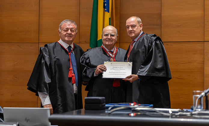 Foto de três homens, todos desembargadores, de vestindo togas pretas, sendo que o do meio porta no peito uma medalha com a qual foi homenageado e segura um diploma junto com homem posicionado à direita