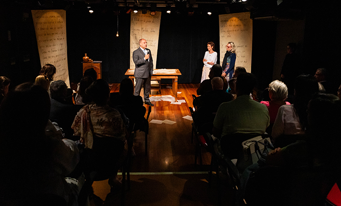 Fotografia mostra sala de espetáculo na penumbra tendo ao palco um homem de terno, o presidente do TJRJ, desembargador Ricardo Couto de Castro, e duas mulheres, a desembargadora Cristina Gaulia e a atriz Ana Barroso