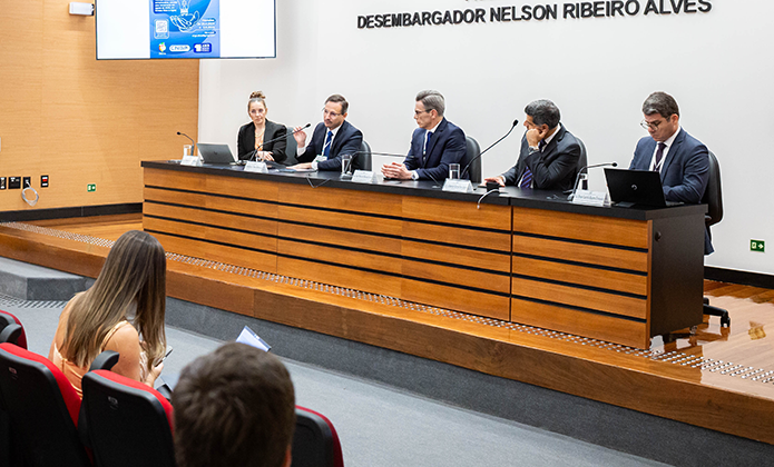 Foto mostra uma mulher de casaco preto e blusa em tom bege e quatro homens vestidos com ternos escuros. Todos estão sentados em mesa de auditório, um pouco elevada em relação à plateia, e têm microfones à frente, pois estão palestrando e debatendo com a plateia, sentada à frente, como mostra a imagem., composta ainda por um telão que aparece ao fundo, à esquerda. A cena se passa em auditório de paredes claras e piso cinza