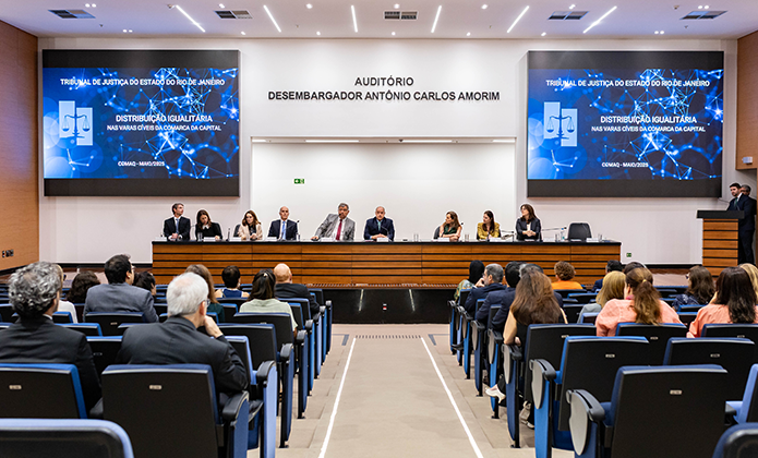 Foto mostra evento em auditório com oito pessoas na mesa principal, sendo quatro homens e cinco mulheres, falando para plateia acomodada em cadeiras azuis de fundo preto. O ambiente é claro