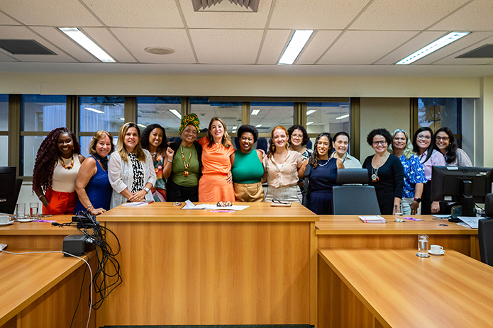 Mulheres vestidas com diferentes roupas e cabelos posam para uma foto, de pé, sorrindo, em sala de reunião