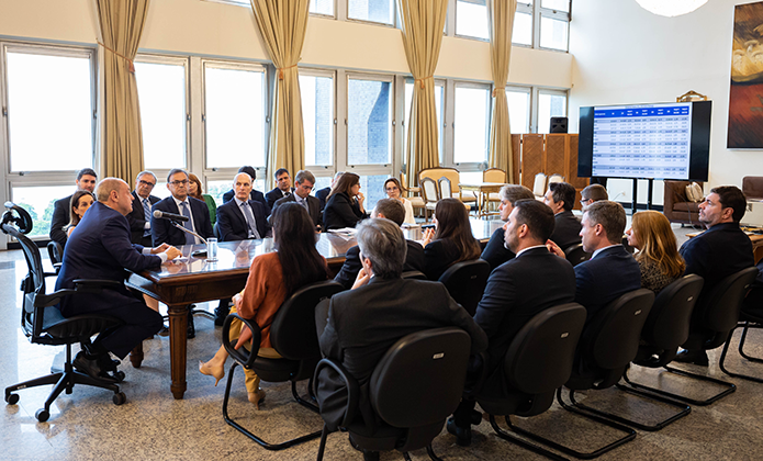Sentado à cabeceira de uma grande mesa, em salão cheio de janelas envidraçadas por onde entra luz natural,  presidente do TJRJ, Ricardo Couto, de terno escuro, comanda reunião com seus auxiliares diretos, homens e mulheres, sentados nos dois lados da mesa, em cadeiras enfileiradas de forma paralela. Ao fundo, um painel mostra números e dados expostos no encontro