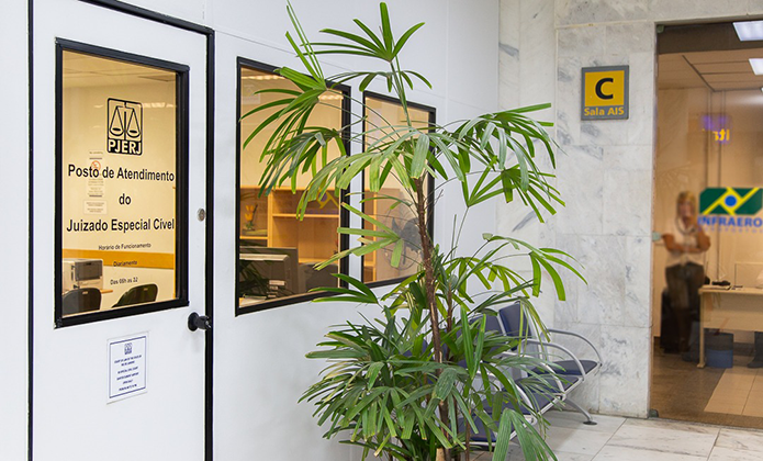 Foto da fachada da sala do Juizado Especial Cível no aeroporto mostrando a porta com parcial visão por conta de uso de  vidro, bem como janelas de vidro lacradas apenas para oferecer a visão do interior do recinto. à frente da sala há bancos para espera de atendimento e uma pequena palmeira 