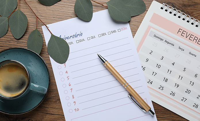 #ParaTodosVerem: Em uma superfície de madeira, há uma xícara de café com espuma na borda, um bloco de notas destinado a registrar aniversários com espaços em branco e números ao lado, e uma caneta dourada apoiada sobre ele. À direita, um calendário de mesa espiral mostra o mês de fevereiro. Algumas folhas verdes decorativas ocupam a parte superior da imagem.