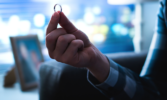 #ParaTodosVerem: Uma mão masculina segura delicadamente uma aliança entre o polegar e o indicador, em ambiente interno de luz suave. O fundo está desfocado, revelando apenas manchas de luz coloridas e, mais ao canto, a silhueta vaga de um porta-retratos.