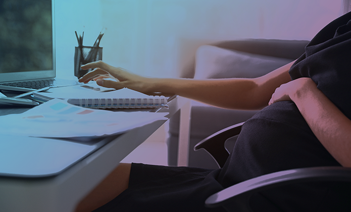 #ParaTodosVerem: Pessoa grávida sentada em cadeira de escritório diante de uma mesa com notebook aberto. Uma das mãos está sobre a barriga, enquanto a outra utiliza o computador. Sobre a mesa há papéis, um caderno espiralado e um porta-canetas. A cena tem iluminação suave em tons azulados e rosados, sugerindo ambiente de trabalho doméstico ou escritório.