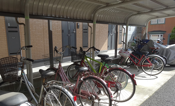 #ParaTodosVerem: Fotografia em ambiente externo mostra um bicicletário coberto por estrutura metálica de telhado curvo. Sob a cobertura, há várias bicicletas estacionadas lado a lado. Ao fundo, vê-se a fachada de um prédio residencial em tons claros.