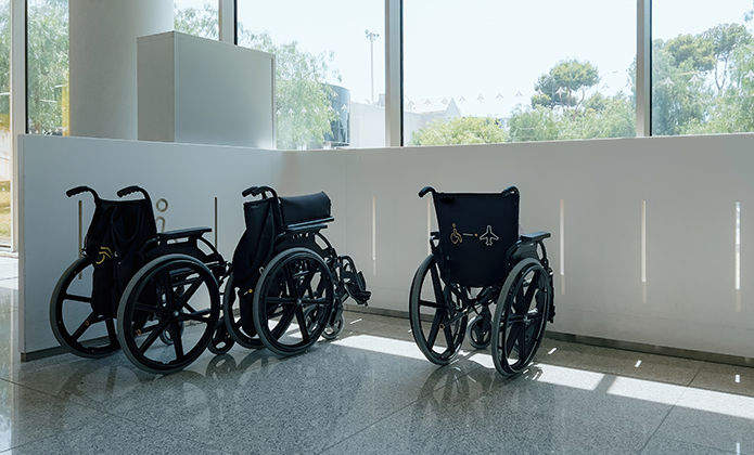 #ParaTodosVerem: Três cadeiras de rodas pretas estão alinhadas próximas a uma parede branca, em um saguão iluminado por janelas amplas. O piso de granito reflete discretamente as cadeiras. Árvores aparecem do lado de fora.