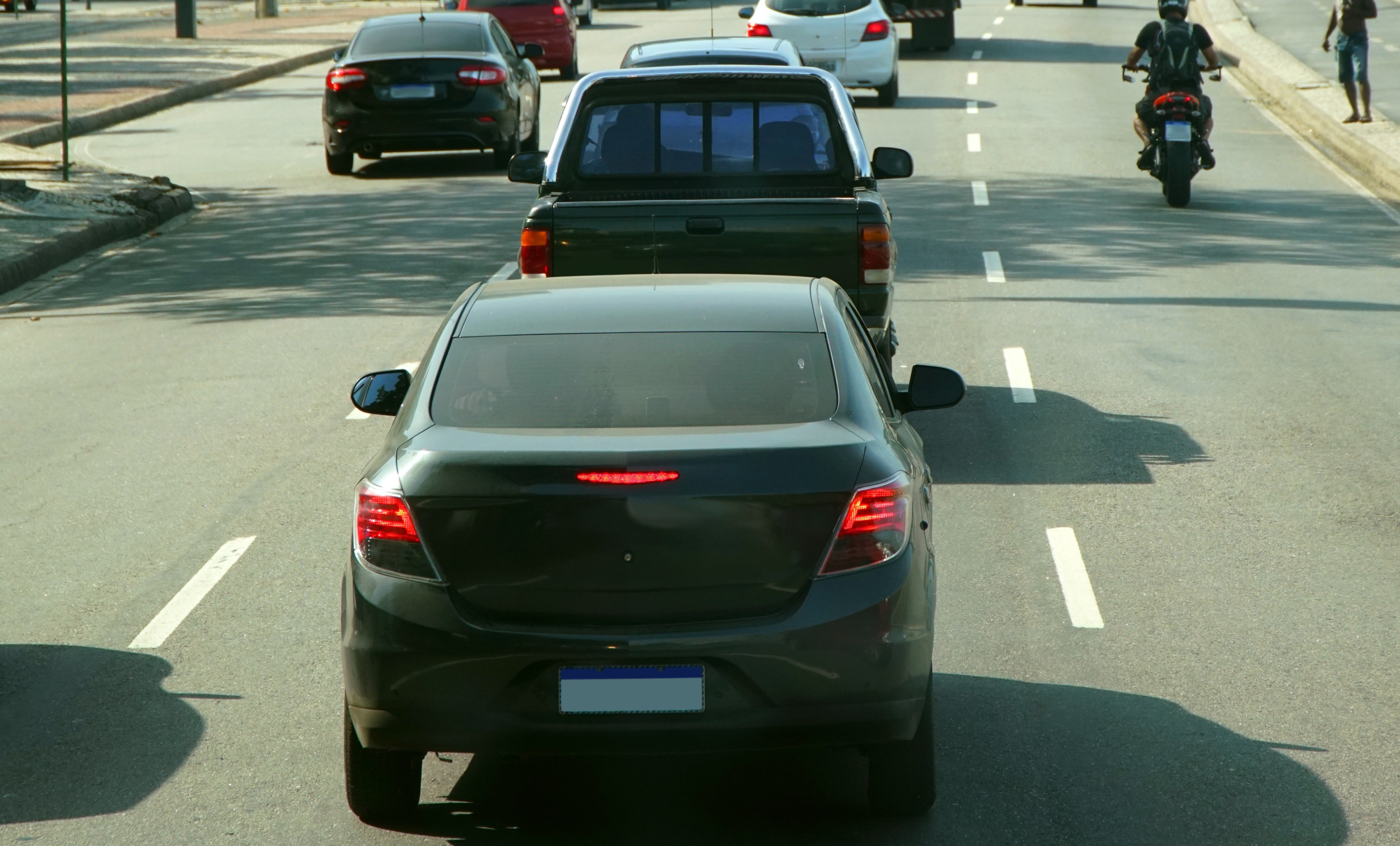 #ParaTodosVerem: Vista traseira de um carro parado no trânsito, seguido por uma picape e outros veículos ao longe. À direita, um motociclista passa entre as faixas, e um pedestre caminha pela calçada. A luz forte realça as marcas brancas da pista e as sombras que se estendem pelo chão.