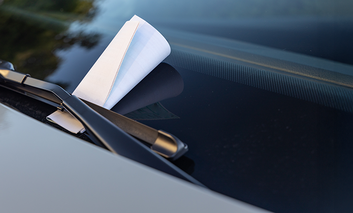 #ParaTodosVerem: Imagem mostra o para-brisa de um carro visto de perto. Um papel branco dobrado, parecido com um aviso ou multa, está preso sob a palheta do limpador de para-brisa. O vidro reflete o céu e árvores, indicando que o carro está estacionado ao ar livre, durante o dia.