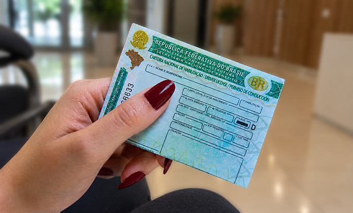 Fotografia em close mostra uma mão segurando uma Carteira Nacional de Habilitação (CNH) do Brasil. As unhas estão pintadas de vermelho escuro. O documento ocupa o centro da imagem e está levemente inclinado, com parte das informações visíveis. Ao fundo, em desfoque, há um ambiente interno com piso claro e móveis, destacando o documento em primeiro plano.