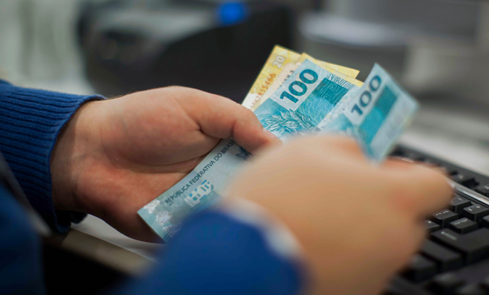 #ParaTodosVerem: Imagem em close mostra mãos segurando cédulas de cem reais diante de um teclado de computador, sugerindo operação financeira ou pagamento em dinheiro.