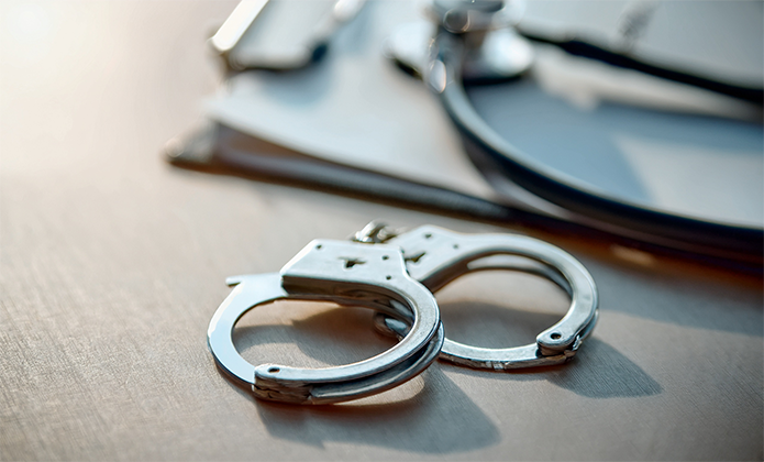 #ParaTodosVerem: Um par de algemas metálicas repousa sobre uma mesa clara, com um estetoscópio e uma prancheta desfocados ao fundo.