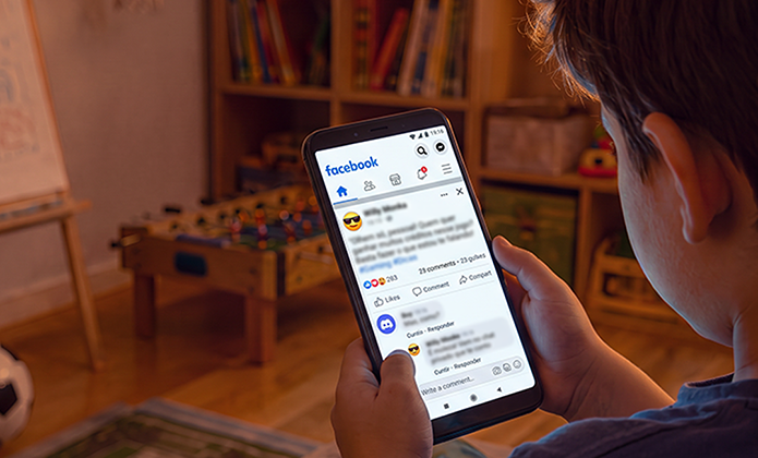 Imagem mostra uma pessoa segurando um smartphone e visualizando a página inicial do Facebook. A tela exibe publicações, ícones de interação e opções de navegação da rede social. Ao fundo, desfocado, há um ambiente interno com estante de livros, mesa de jogos e mobiliário residencial. A iluminação é suave e quente, destacando o aparelho celular em primeiro plano e remetendo ao uso de redes sociais em ambiente doméstico