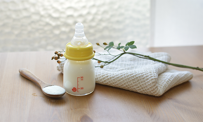 #ParaTodosVerem: Um frasco pequeno de mamadeira com tampa e bico amarelos está cheio de leite até a marca de 50 ml. Ele está apoiado sobre uma superfície de madeira clara. Ao lado, há uma colher de madeira com um pouco de leite em pó e um pano dobrado de tecido macio, também claro. Sobre o pano, repousa um galho fino com pequenas folhas verdes e botões secos. 