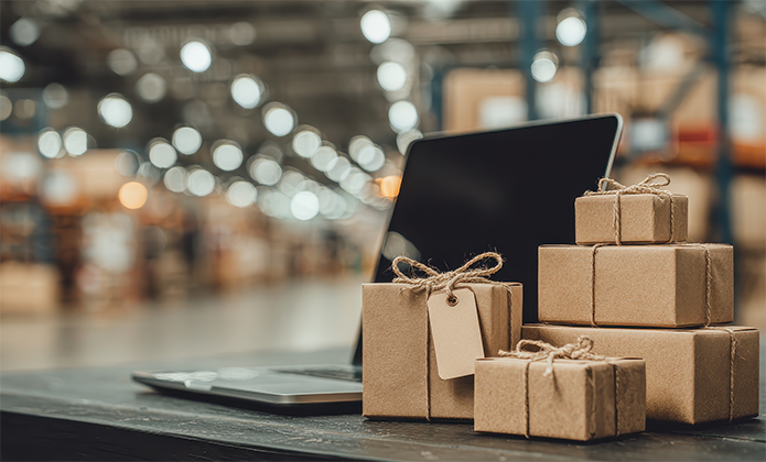 #ParaTodosVerem: Uma pilha de caixas de papel pardo amarradas com barbante está sobre uma mesa, ao lado de um notebook aberto. Ao fundo, desfocado, vê-se o interior de um grande galpão iluminado, com prateleiras e luzes pendentes, sugerindo um ambiente de armazém ou centro de distribuição.