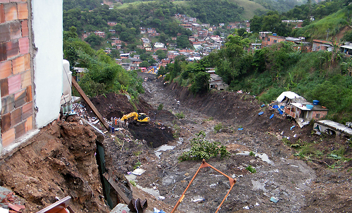 #ParaTodosVerem: Vista ampla de uma área de encosta marcada por deslizamento de terra ou erosão. Em primeiro plano, aparecem solo exposto, entulho, vegetação danificada e parte de construções próximas à borda do terreno. Ao centro, há uma máquina amarela de escavação em área de terra revolvida. Ao fundo, vê-se uma comunidade espalhada entre morros verdes, sugerindo cenário de risco geológico, desastre ambiental ou intervenção urbana em região habitada.