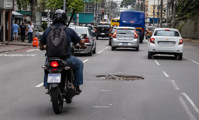 Fotografia de via urbana movimentada. Em primeiro plano, uma motocicleta trafega pela faixa da esquerda. No centro da pista, há um grande buraco no asfalto. Mais à frente, carros e um ônibus circulam pela avenida. Ao fundo, aparecem imóveis, árvores e outros