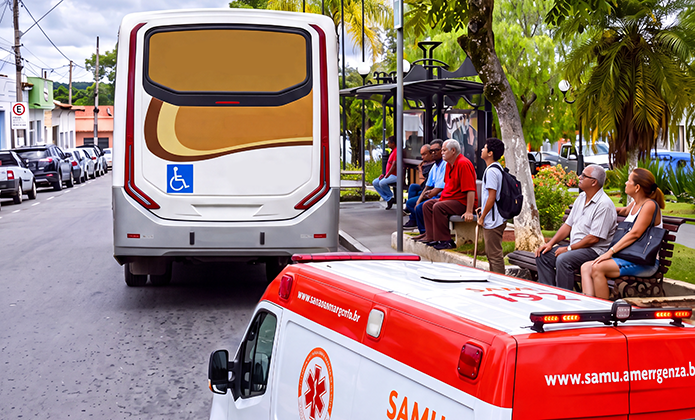 Imagem mostra a traseira de um ônibus urbano parado em um ponto de embarque, com símbolo de acessibilidade para cadeirantes na parte inferior. Ao lado do veículo, várias pessoas aguardam no abrigo do ponto e em bancos instalados na calçada arborizada. Em primeiro plano, aparece parcialmente uma ambulância do SAMU estacionada na via. Ao fundo, são vistos árvores, veículos e edificações urbanas. 
