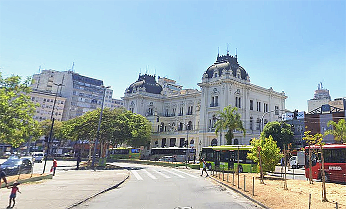 ParaTodosVerem: Foto mostra prédio histórico de fachada branca com cúpulas, em área urbana ensolarada; ao redor há árvores, ônibus circulando e prédios modernos ao fundo.