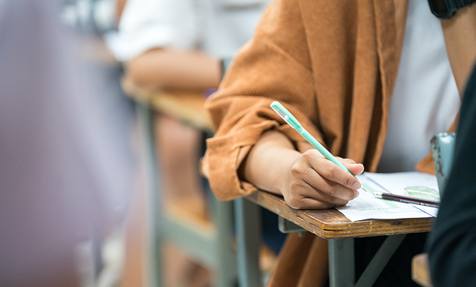 #ParaTodosVerem: Imagem mostra uma pessoa sentada em carteira escolar escrevendo numa folha com lápis, enquanto outras pessoas aparecem desfocadas ao fundo, sugerindo ambiente de sala de aula ou prova.