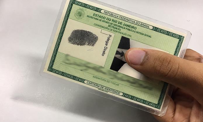 Imagem mostra uma carteira de identidade do Estado do Rio de Janeiro sendo segurada por uma mão. O documento está plastificado e aparece inclinado sobre uma superfície clara. Dados pessoais, fotografia e assinatura estão desfocados, preservando a identificação do titular. No topo do documento, é possível ler “Estado do Rio de Janeiro” e “Carteira de Identidade”.