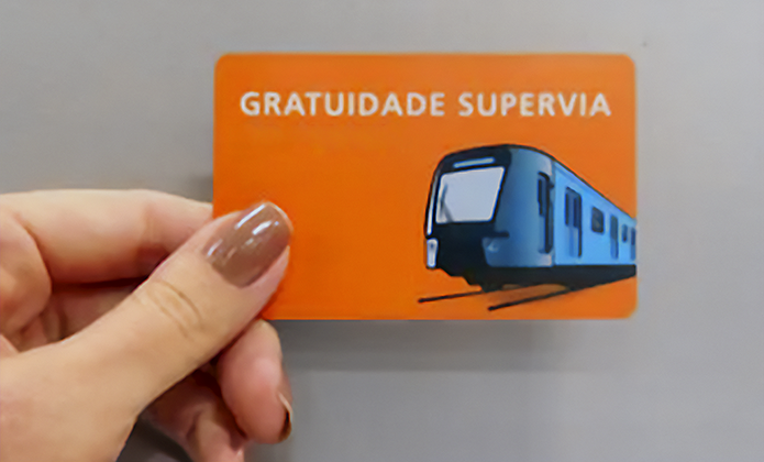 #ParaTodosVerem: Close de uma mão segurando um cartão laranja de transporte público. No cartão, lê-se “GRATUIDADE SUPERVIA” em letras brancas, acompanhado da ilustração de um trem azul sobre trilhos. O fundo é cinza e desfocado, destacando o cartão, que remete ao benefício de gratuidade no transporte ferroviário.