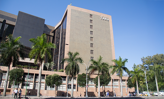Fotografia colorida da fachada do Tribunal de Justiça do Estado do Rio de Janeiro (TJRJ) em dia ensolarado. O edifício de grande porte, com revestimento em tons claros e a sigla “TJRJ” próxima ao topo, ocupa a maior parte da imagem. À frente da construção há diversas palmeiras alinhadas, árvores menores, postes de iluminação e uma área de circulação com pedestres. O céu está azul e sem nuvens, destacando a arquitetura do prédio e a vegetação ao redor.