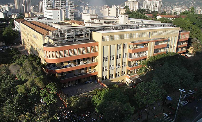 Legenda: #ParaTodosVerem: Vista aérea de um grande edifício urbano de vários andares, em tons de bege e terracota, cercado por árvores densas. O prédio ocupa quase todo o quarteirão e possui varandas curvas e áreas abertas. Ao fundo, aparecem diversos prédios residenciais e comerciais espalhados pela cidade, em meio a áreas verdes e morros. A imagem foi feita durante o dia, com iluminação natural e sombras projetadas pelas árvores e construções.
